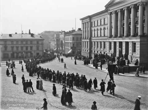 Työläisten muodostama kulkue kulkee Senaatintorin läpi yliopiston päärakennuksen editse. Kulkueen edessä kannatellaan isokokoista viiriä. Väkijoukko seuraa kulkueen etenemistä katujen varsilla.