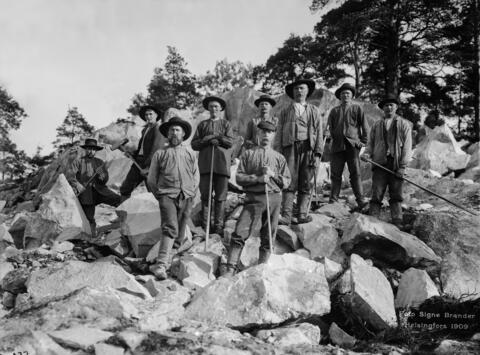 En grupp män bryter sten år 1909. Nio i enkla kläder och hattar klädda män lutar sig mot sina arbetsredskap i mitten av ett stenbrott och poserar för kameran. 