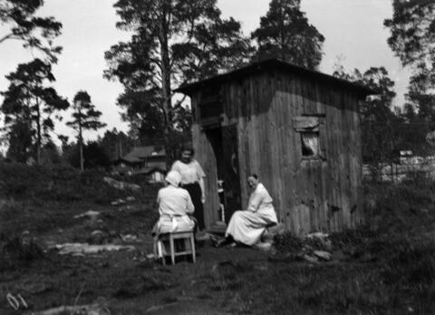 Kolme naista pienen puuhökkelin edustalla. Rakennus on ilmeisesti vanha työkaluvaja, tuskin muutamaa neliömetriä suurempi. Taustalla metsäistä kalliomaisemaa.