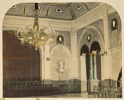 An old photograph of an antique-themed banquet hall. There is a large chandelier and Greek-style columns.