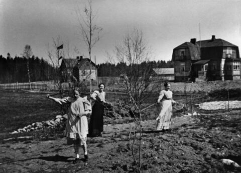 Kuvan etualalla kolme naista kesäpelin tiimellyksessä, taka-alalla kaksi huvilaa. Maassa on lumipälviä ja puut ovat lehdettömiä. 