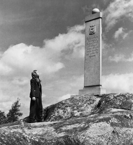 A woman is standing next to a stone column on a rock. The woman is wearing a long overcoat and a veil on her head.