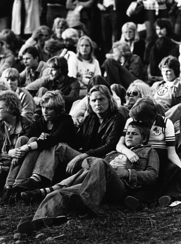 Nuorta yleisöä istuu nurmikolla Kaivopuiston konsertissa. Monilla on pitkät hiukset ja leveälahkeiset housut. Yksi katsoja makaa toisen sylissä.