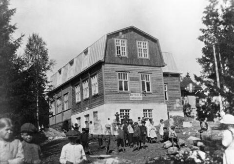 Keskellä koulurakennus, jonka seinässä kyltti Metsola kansakoulu. Etualalla lapsia, pojilla lakit päässä, tytöillä esiliinat.