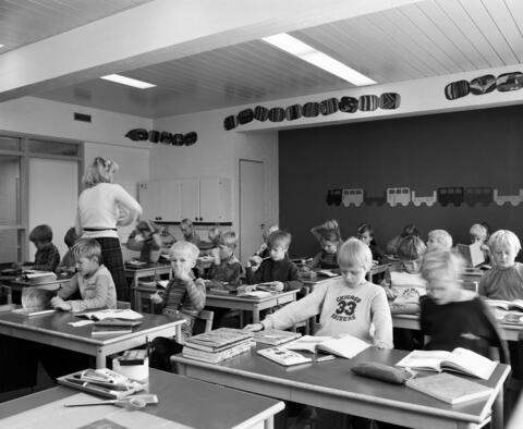 Alakoululaisia oppitunnilla luokassaan. Oppilaiden pöydillä on kirjapinoja, opettaja kiertää luokassa.