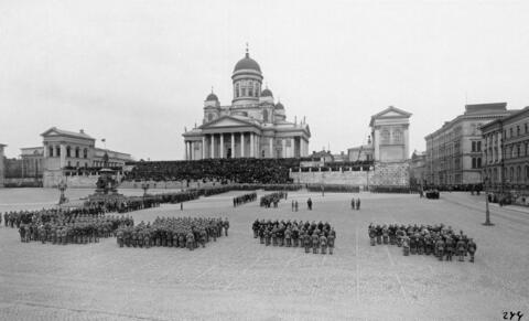 Jääkärit ovat järjestäytyneet paraatimuodostelmiin ympäri Senaatintoria. Ryhmittymien keskelle on kokoontunut oletettavasti jääkäriprikaatin päällystöä. Yleisö on kerääntynyt seuraamaan tapahtumia tuomiokirkon portaille.