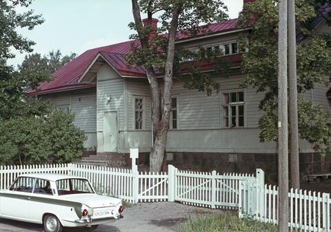 Edustalla näkyy valkoinen 1960-luvun Ford Cortina GT -auto. Taustalla näkyy valkoinen puurakennus, jonka harjakatto on punainen ja sieltä nousee kaksi savupiippua. Rakennusta ympäröi valkoinen matalahko aita ja pensaita ja puita.