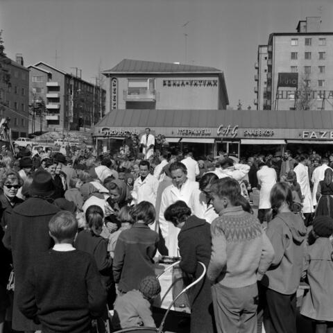En stor skara människor har samlats framför köpcentret. Representanter för Lions club skiljer sig från mängden med sina vita jackor. En av dem håller tal genom att tala i mikrofon på ett podium. I bakgrunden syns våningshus. 