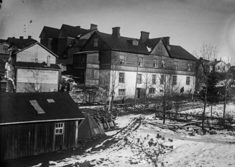 Puisia monikerroksisia asuinrakennuksia Pasilassa, talvisessa maisemassa 1900-luvun alussa. Kuvan etualan rakennuksessa on harjakatto ja kolme kerrosta. Taloja on rakennettu tiiviisti vierekkäin.