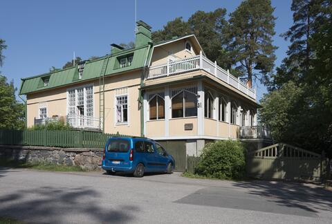 Vaaleankeltainen puuhuvila, jossa on iso veranta ja parveke sekä paljon pieniruutuisia ikkunoita. Talon edessä on pysäköity auto.