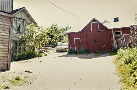 En gårdsplan i Trä-Böle. Till vänster ett bostadshus, mitt på bilden en klarröd lada, som tydligen fungerar som garage.