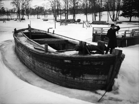 Proomu Hoppet talvella. Taustalla rantaa, jossa on maassa rumpuputkia. Ragnar Nyberg seisoo proomun takana jäällä, laiturin vieressä