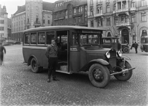 Rautatientorilla Fennian talon edessä linja-auto, jonka kuljettaja lakki päässä ja nahkasaappaat jalassa seisoo auton vieressä.