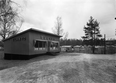 En liten träbyggnad med pulpettak. På båda väggar texten ”ELANTO”. Byggnaden har stora fönster på framsidan mot sandfältet. I bakgrunden syns koloniträngårdsstugor.