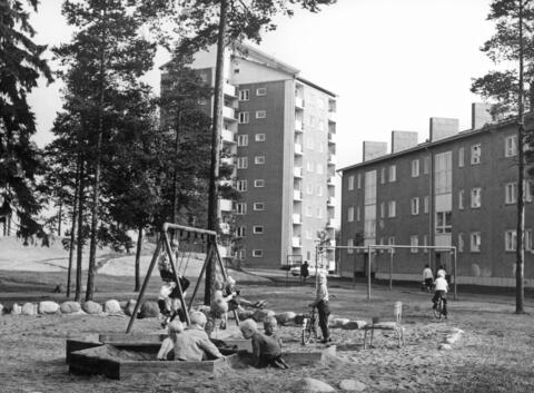 I förgrunden leker barn i en lekpark. En del av barnen är i sandlådan, några gungar och några har cyklar. I bakgrunden syns våningshus och träd. 