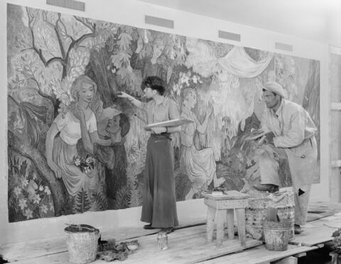 A large fresco with a slim woman standing in front of it, painting it. A male assistant is standing nearby, leaning his foot on a large paint can.
