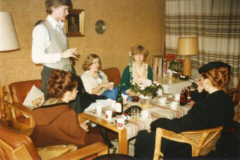 En grupp festklädda gymnasister sitter avslappnat i ett vardagsrum med drinkar. 