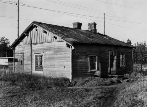 Puinen yksikerroksinen, kaksipiippuinen rakennus hallitsee kuvaa. Rakennuksessa on harjakatto ja se on huonokuntoinen. Rakennuksen portailta lähtee polku ja vasemmanpuoleiselta seinustalta ristikkoaita.
