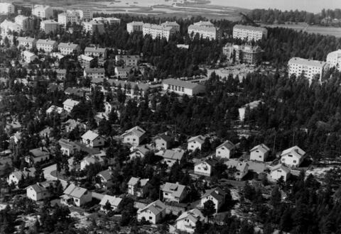 I förgrunden syns ett småhusområde. Baktill syns våningshus av olika storlekar. Området är grönskande. Olika slags träd växer mellan husen.