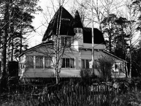 En ljus villabyggnad av trä domineras av ett stort torn som har står sida vid sida med ett mindre torn med smalare brantare spets. Byggnaden har 3–4 våningar. Gården ser rätt ovårdad ut.
