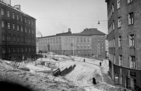 Kerrostaloja, maassa lunta. Muutama ihminen kävelee kadulla.