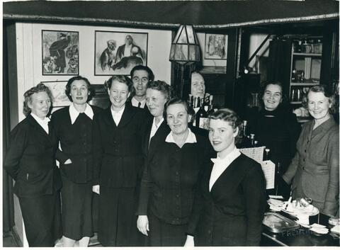 Åtta kvinnor och tre män står bakom eller framför en serveringsdisk i en restaurang. Kvinnorna bär mörka dräkter och vita skjortor. På väggen är konstverk.