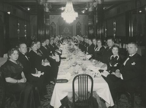 Pitkän katetun pöydän ääressä istuu juhlapukuisia miehiä ja naisia. Katossa on kristallikruunut.