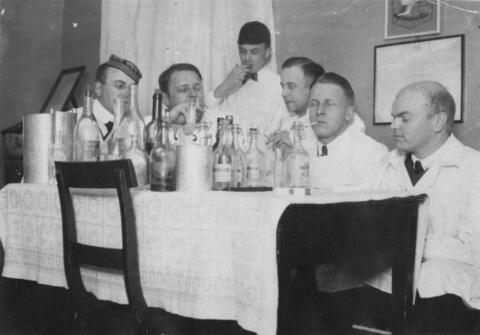 Kuusi valkotakkista miestä istuu pöydän ääressä, joka on täynnä viinapulloja. Neljä miehistä tupakoi.