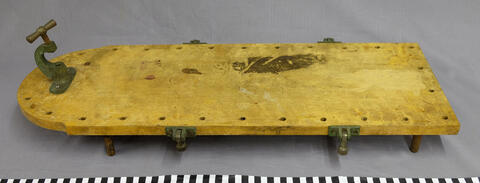 Vaaleaa puuta oleva leikkuulauta, jonka alla on matalat jalat ja metallikiinnikkeet sivuilla. Sitä on pidetty tavallisen pöydän päällä. 