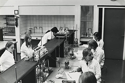 Seitsemän valkoisiin takkeihin pukeutunutta opiskelijaa istuu laboratorion työpöytien ääressä. Pöydillä näkyy laboratoriovälineitä.