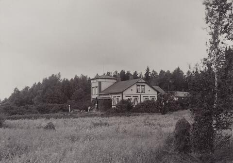 Ett svartvitt fotografi föreställer ett åkerlandskap med en tvåvåningars byggnadshelhet i mitten. 