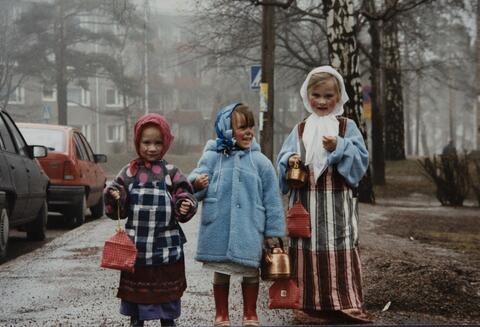 Tre barn utklädda till påskhäxor står på en trottoar med kopparkaffepanna i handen. Längs med gatan står bilar parkerade.