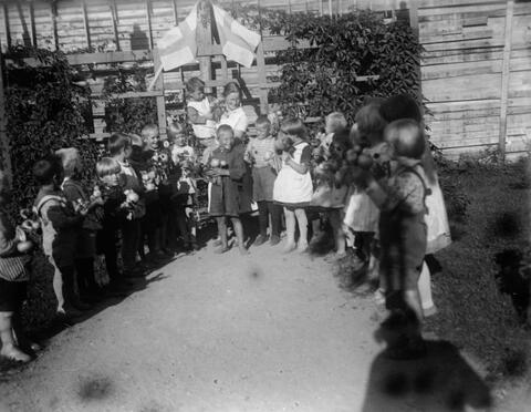 Lapsia ja muutama aikuinen kokoontuneena puoliympyrän muotoon lastentarhan pihalle. Taustalla on kaksi Suomen lippua ja köynnöskasveja.
