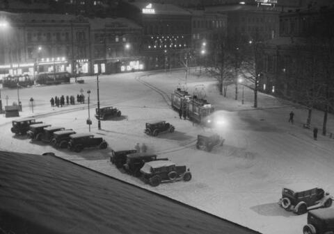 Katolta otetussa valokuvassa on luminen maisema Ruotsalaisen teatterin vierestä. Ihmisiä seisoo pysäkillä ja raitiovaunu on tulossa kääntöraiteelle. Autoja on pysäköity aukiolle.