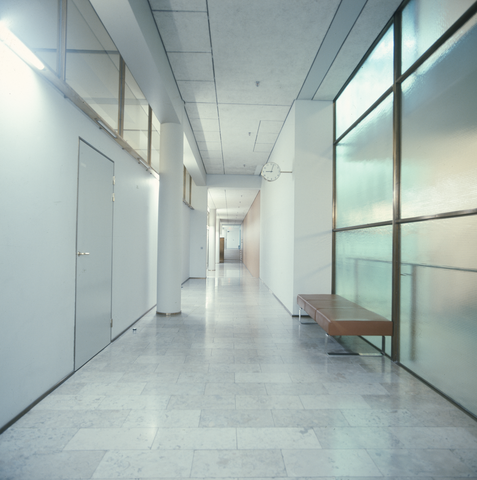 A photo of a corridor with white walls, and a column and a door on the left. There is a glass wall with a leather seat in front of it on the right.