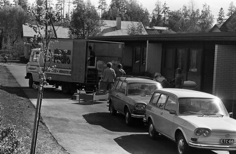 Rivitalon pihalla on muuttoauto, johon lastataan huonekaluja. Lisäksi etualalla on kaksi autoa.