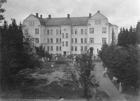 Pihatie ja puisto, jossa on ihmisiä. Taustalla kohoaa mansardikattoinen moniosainen kivirakennus.