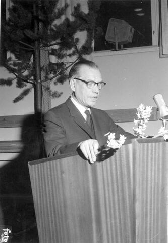 En kostymklädd man håller tal i en talarstol av trä.