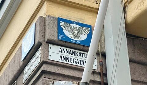 Rakennuksen kulma, johon on kiinnitetty korttelikilpi ja kadunnimikilvet. Korttelikilvessä lukee Pöllö Ugglan kortteli kvarteret 89, ja siinä on piirretty pöllö. 