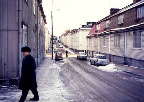 Näkymä Vanajantie 19:n kohdalta Vallilantielle päin. Oikealla Vanajantie 16-18, 14, 12. Mies kävelee Vanajantie 19:n pihalle. Kuvassa autot Ford Anglia, Fiat 600 ja Moskvitš Elite. Teiden varsilla näkyy hieman lunta.