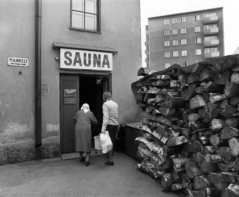 Kaksi henkilöä astumassa sisään yleiseen saunaan. Oven vieressä on parin metrin korkuinen pino paksuja polttopuita. Saunan ovessa kyltit, joissa aukioloajat ja ilmoitus mahdollisuudesta mankelin vuokraamiseen.