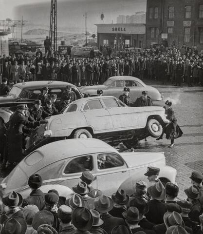 Suuri väkijoukko kadulla. Poliisit ovat nostamassa autoa takaisin renkailleen. 