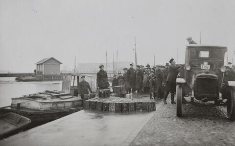 Satama-altaassa on vene, jossa seisoo mies. Altaan reunalla on kymmeniä miehiä, kuorma-auto ja kymmeniä metallikanistereita. 