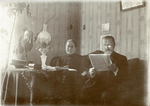 Mies ja nainen istuvat nojatuoleissa. Nainen katsoo kuvaajaan, mies katsoo kädessään olevaa sanomalehteä. Pöydällä on kirjoja ja öljylamppu. Kummallakin on silmälasit.