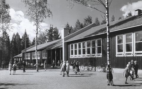 Lapsia ja polkupyöriä yksikerroksisen harjakattoisen koulurakennuksen pihalla.
