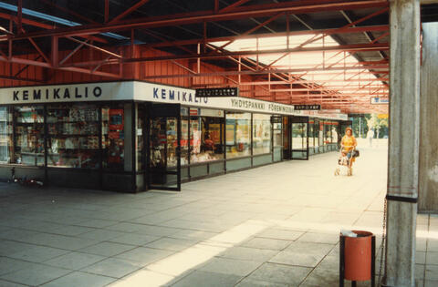Etualalla on harmaita betonilaattoja, vasemmalla liikerakennus, jossa lukee KEMIKALIO ja YHDYSPANKKI. Oranssiin mekkoon pukeutunut nainen työntää lastenvaunuja oikealla.
