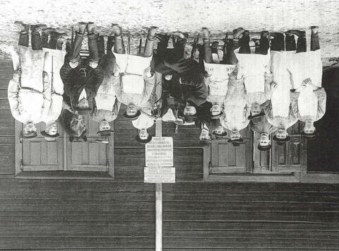 Viisitoista miestä, joilla on hattu päässä. Lähes kaikilla on valkoinen esiliina. Keskellä on hevonen ja lehmä. Taustalla on hirsiseinä.