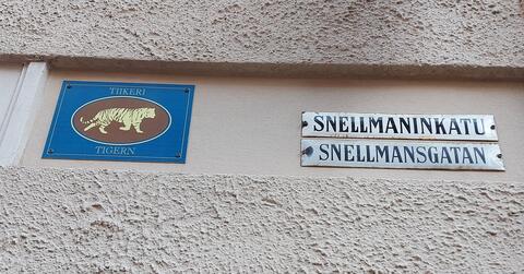 Putsad husvägg på vilken man fäst en skylt med en ritad tiger och texten TIIKERI TIGERN. Bredvid den gatuskyltarna SNELLMANINKATU SNELLMANSGATAN.