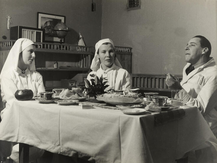 Etualalla on katettu kahvipöytä, vasemmalla istuu kaksi valkoisiin työasuihin pukeutunutta sairaanhoitajaa, oikealla istuu lääkärintakkiin pukeutunut mies tupakoimassa. 