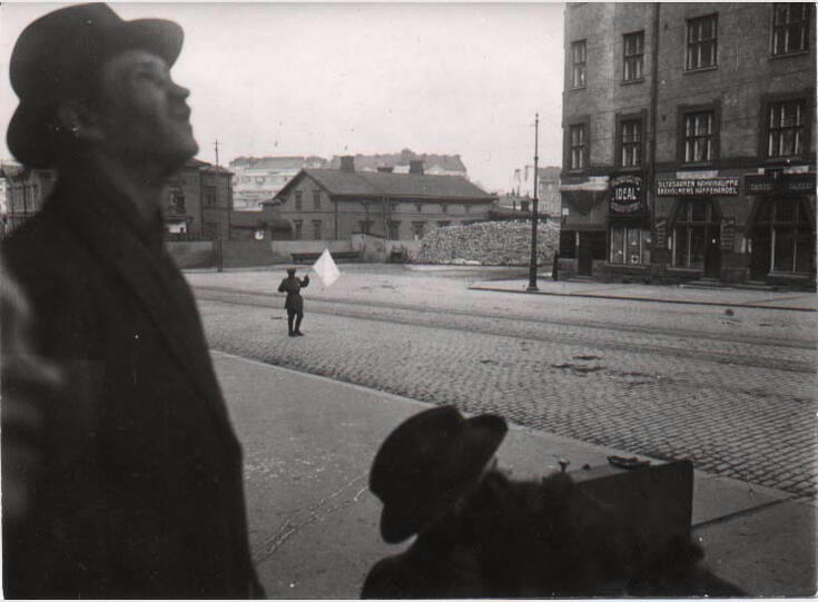 En ung man bär en vit flagga på Broholmsgatan. I förgrunden två män som tittar snett uppåt. 